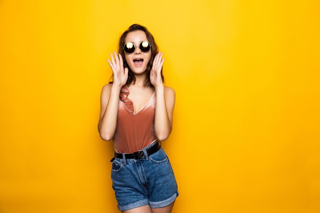 Sonnenbrille der jungen Frau, die weg mit überraschtem Lächeln lokalisiert auf gelber Wand schaut.