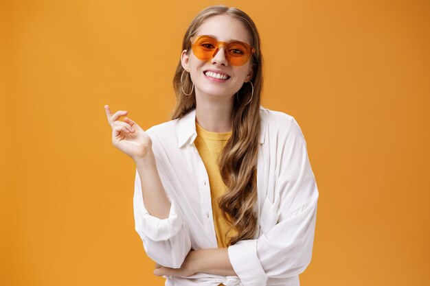 Sonnenbrille im passenden Stil. Porträt einer selbstbewussten und sorglos gut aussehenden Modebloggerin in Brillen und weißem T-Shirt, die mit erhobener Hand gestikuliert und fröhlich in die Kamera lächelt.