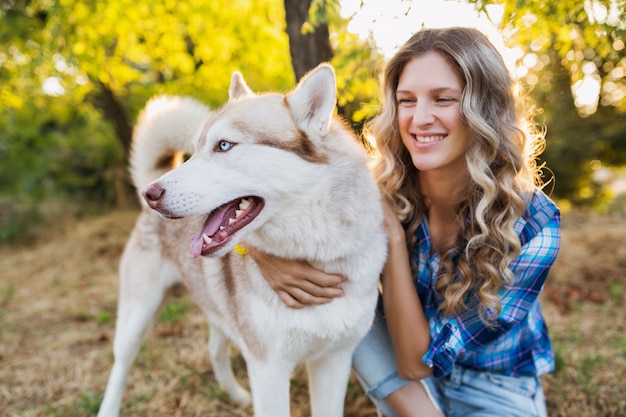 Sonnige junge stilvolle hübsche lächelnde glückliche blonde Frau, die mit Hund husky Rasse im Park am sonnigen Sommertag spielt