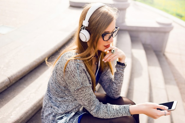 Sonniges Bild des Sommerlebensstils der hübschen jungen blonden Frau, die Musik durch Kopfhörer hört, Handy hält, auf der Straße sitzt, träumt. Trage ein stylisches Frühlingsoutfit.