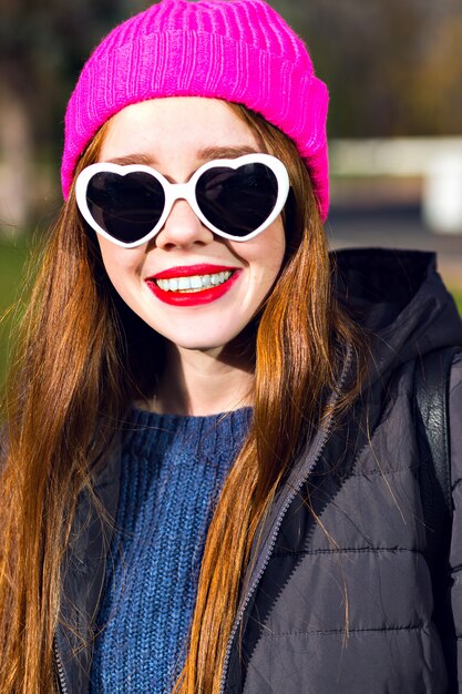 Sonniges Frühlingsporträt der glücklichen fröhlichen lächelnden Ingwerfrau, die im Park aufwirft, genießen sonnigen Tag, hellen Punk-Hipster-Hut, herzige Sonnenbrille, rote Lippen, warmen Parka, positive Stimmung.