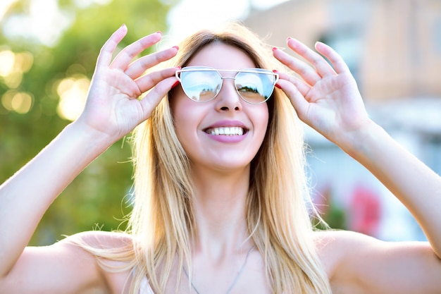 Sonniges Frühlingssommerporträt der glücklichen blonden Frau genießen schönen warmen Tag lächelnd und schließen ihre Augen, tragen stilvolle trendige Brille, positive Stimmung.