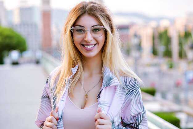 Sonniges Porträt der glückseligen glückseligen jungen Frau des Glaubens flauschige blonde Haare, Frühlingszeit, tragen klare Sonnenbrille und tropische Druckjacke, Reisestimmung.