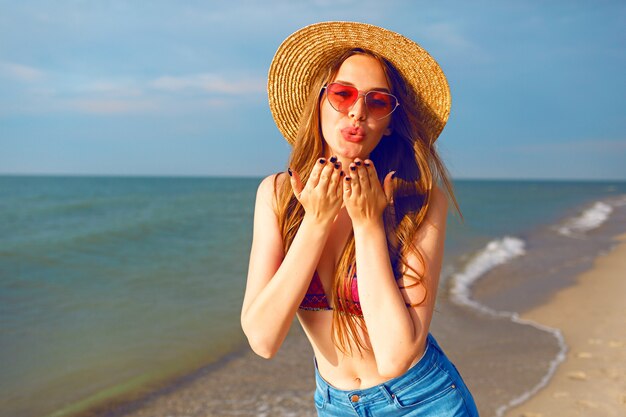 Sonniges Sommerporträt des blonden hübschen Mädchens, das Spaß am Strand in der Nähe des Meeres hat, Zunge zeigt und verrückt wird, Hutsonnenbrille tragend