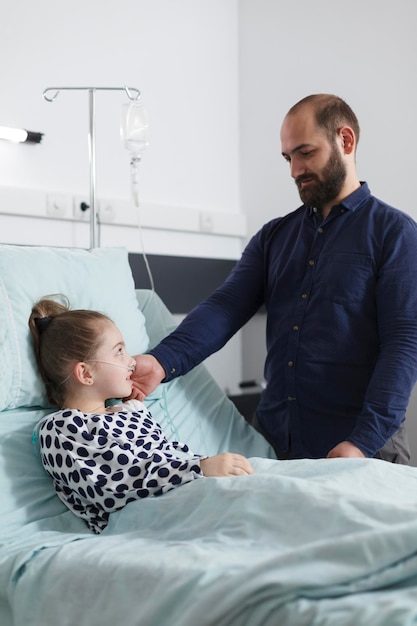 Sorgfältiger unterstützender Vater streichelt kranke kleine Tochter, während er im Krankenhausbett liegt.