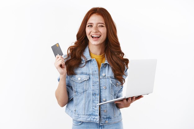 Sorglos entspanntes rothaariges Mädchen, das versichert, dass alles in Ordnung ist, solange sie eine Einzahlung hat, die eine schwarze Kreditkarte zeigt, die einen frechen Laptop hält, der zufrieden mit einem Geschenk für einen Freund im Internet-Shop lächelt