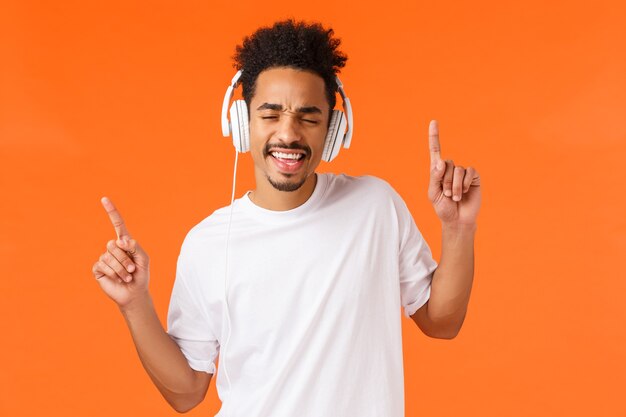 Sorglos glücklicher und erleichterter afroamerikanischer gutaussehender Hipster-Typ, der Musik in Kopfhörern hört, tanzt und Hände in den Rhythmus schüttelt, geschlossene Augen, die in Kopfhörern mitsingen, orangefarbener Hintergrund