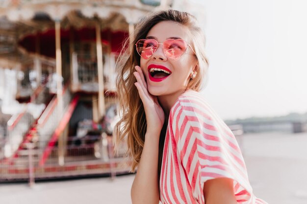 Sorglos schöne Dame lächelt auf Straßenhintergrund und berührt ihre Wange Romantisches weibliches Model mit rosa Sonnenbrille im Vergnügungspark