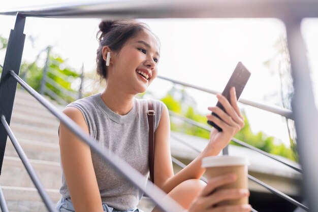 Sorglose digitale Nomadin asiatische Frau hält Kaffeetasse in der Hand, während sie mit dem Smartphone mit entfernten Kunden chattet, während sie auf der Treppe des öffentlichen Parks in der Stadt sitzt und modernen Lebensstil arbeitet