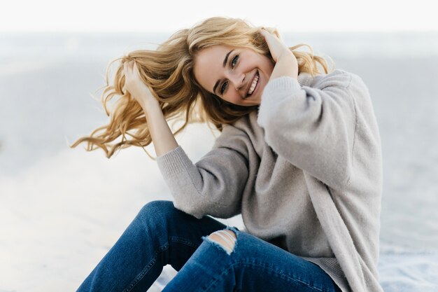 Sorglose Frau im weichen Pullover, die ihr Haar im Strand berührt. Außenporträt der gewinnenden kaukasischen Frau, die am Strand im Herbst ruht.