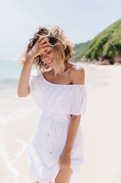 Sorglose junge Dame im Sommerkleid, die ihr welliges Haar berührt. Foto im Freien des glücklichen weiblichen Modells, das am Strand ruht.