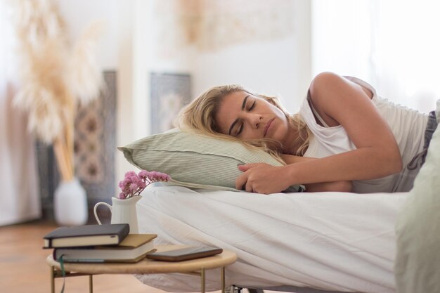 Sorglose kaukasische Frau, die morgens auf ihrer Seite im Bett schläft. Mittlere Aufnahme einer schönen Frau im Schlafanzug, die sich in einem gemütlichen Schlafzimmer mit Nachttisch entspannt. Traum, Schlafkonzept