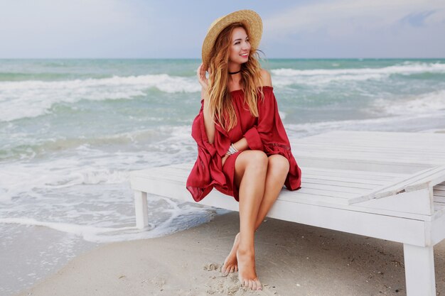 Sorglose liebenswerte Frau im stilvollen Strohhut und im roten Strandkleid, die nahe dem Ozean sitzen. Wochenenden genießen.