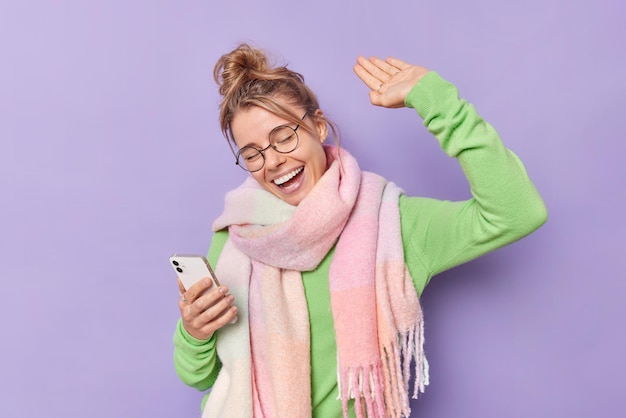 Sorglose, optimistische, hellhaarige Frau hält Handy-Tänze im Rhythmus ihrer Lieblingsmusik, trägt eine runde Brille, grüner Pullover, warmer Schal, lacht glücklich isoliert über violettem Hintergrund, hat Spaß