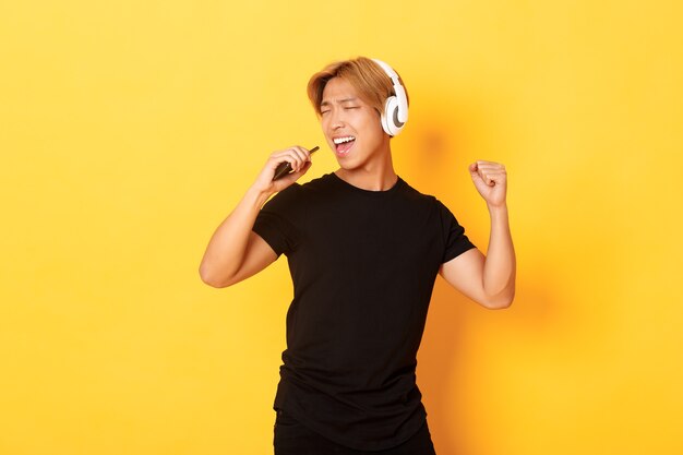 Sorgloser hübscher asiatischer Kerl in den Kopfhörern, Karaoke-App spielend, in Handymikrofon singend, stehende gelbe Wand