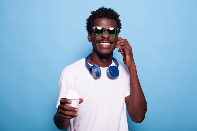 Sorgloser Mann, der mit dem Smartphone telefoniert, während er Sonnenbrillen und Kopfhörer trägt. Junger Erwachsener, der eine Tasse Kaffee in der Hand hält und sich beim Chatten auf dem Gerät fröhlich fühlt