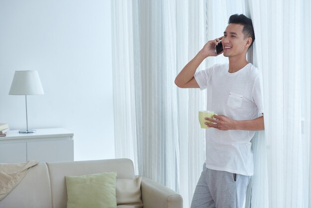 Sorgloser Mann mit der Tasse Tee, der auf seinem Wohnzimmerfenster beim Machen eines Telefonanrufs sich lehnt