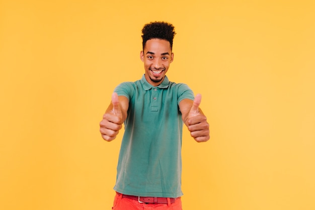 Sorgloser schwarzer Mann im lässigen grünen T-Shirt lächelnd. Innenfoto des emotionalen afrikanischen Kerls, der mit Daumen hoch aufwirft.