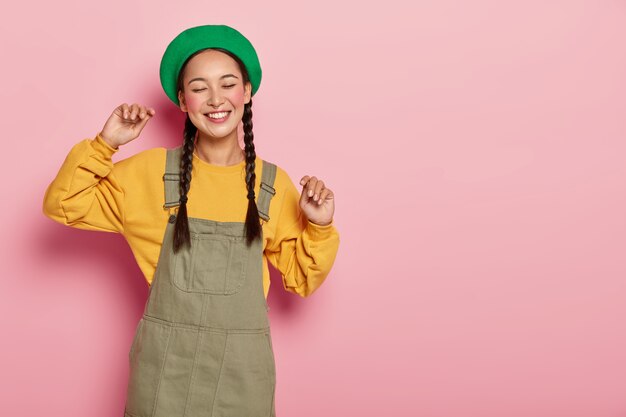 Sorgloses asiatisches Mädchen bewegt sich im Rhythmus der Musik, hält die Arme hoch, trägt grüne Baskenmütze, gelbes Sweatshirt und Sarafan