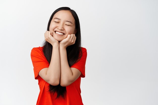 Sorgloses asiatisches Mädchen schließt die Augen und lächelt süß, hält den Kopf an den Händen und träumt im roten T-Shirt auf weißem Hintergrund