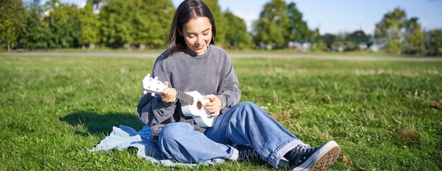 Sorgloses asiatisches Mädchen singt und spielt Ukulele im Park sitzt auf dem Gras und der Musiker entspannt sich auf ihr