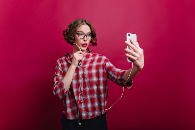 Sorgloses braunhaariges mädchen im karierten freizeithemd, das selfie-innenfoto der fröhlichen jungen dame in den gläsern macht, die mit küssendem gesichtsausdruck aufwerfen und telefon verwenden.