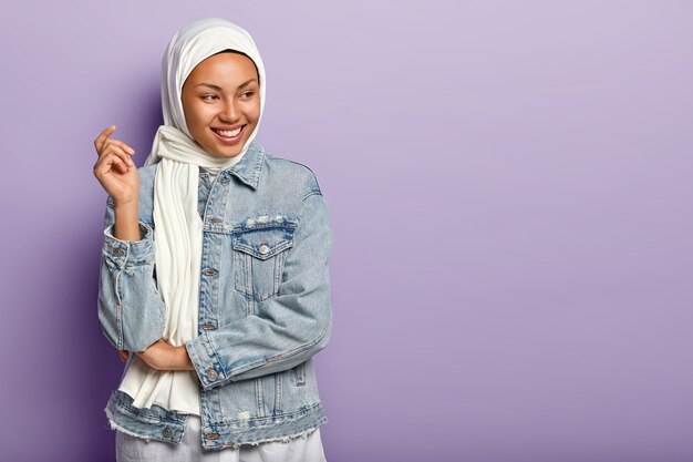 Sorgloses dunkelhäutiges arabisches weibliches Model lacht glücklich, drückt aufrichtige Gefühle aus, lächelt breit, konzentriert beiseite, posiert in Kopfbedeckung und Jeansjacke über lila Wand, Leerzeichen