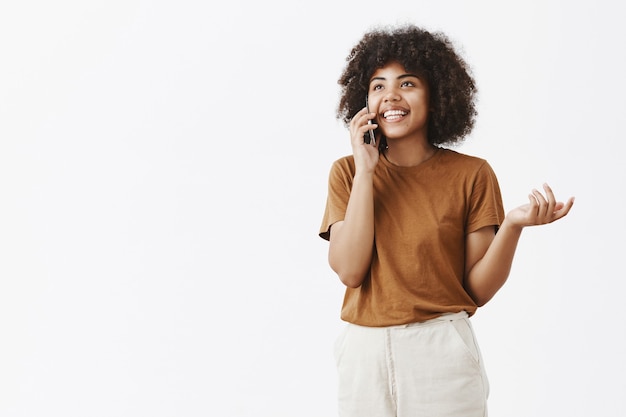 Sorgloses glückliches gut aussehendes und emotionales afroamerikanisches Mädchen mit dem lockigen Haar, das nach oben gestikulierend und lächelnd schaut, während man Handy benutzt