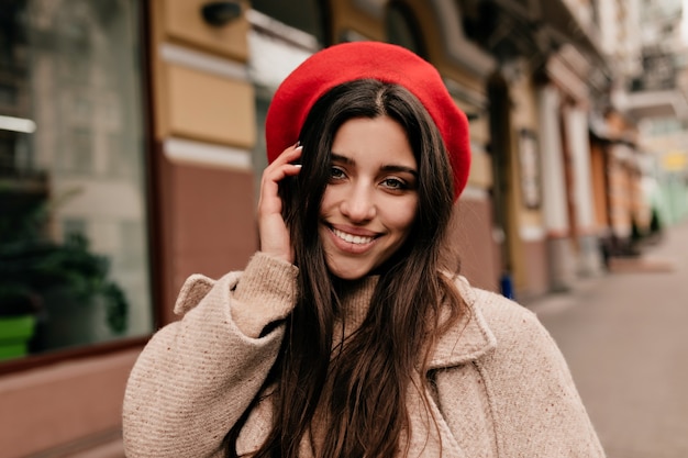 Sorgloses Mädchen im eleganten Hut, der an der Kamera auf Hintergrund der alten Stadt aufwirft. Außenporträt der glückseligen langhaarigen Frau im beigen Mantel, der lachend auf der Straße lacht