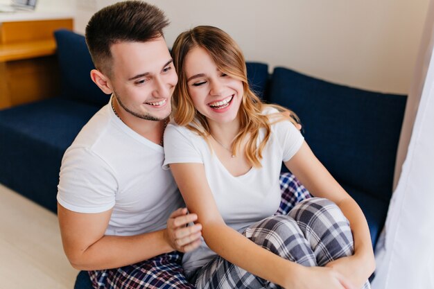 Sorgloses Mädchen im stilvollen Pyjama, das mit ihrem Freund lacht. Glücklicher schwarzhaariger Mann, der Wochenende mit Frau genießt.