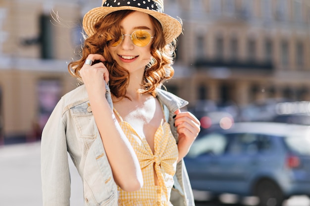 Sorgloses Mädchen in Sonnenbrille und Jeansjacke genießt warmen Frühlingstag. Außenaufnahme der jungen Frau des wunderbaren Ingwers, die während des Gehens sanft lächelt.