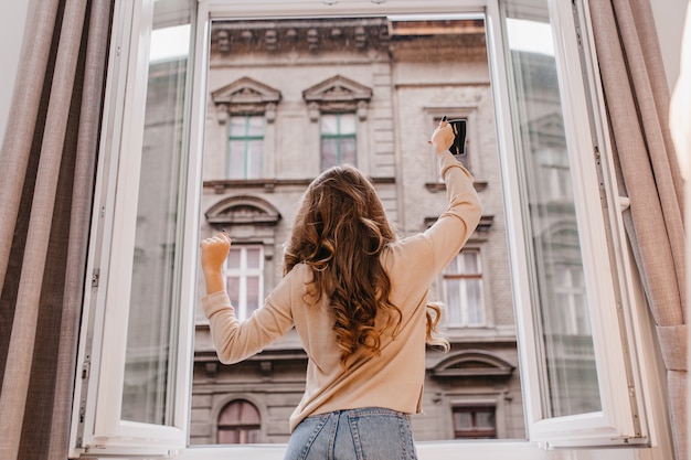 Sorgloses schlankes Mädchen in den Jeans, die neben Fenster mit Tasse Kaffee tanzen