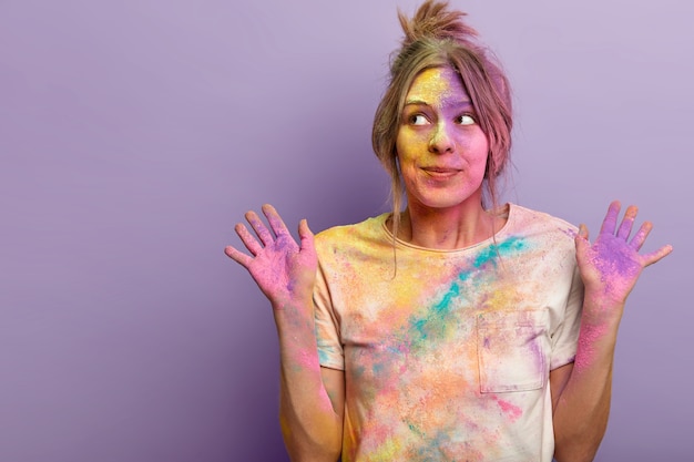 Kostenloses Foto sorgloses schönes weibliches model schaut mit verträumtem neugierigem ausdruck zur seite, hebt mit buntem puder verschmierte handflächen, die an holi-festivalfeiern beteiligt sind, steht über lila wand, leerzeichen