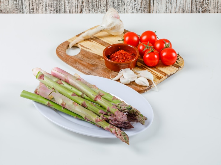 Kostenlose Foto spargel mit tomaten, knoblauch und chilipulver auf schneidebrett in einem weißen teller auf weißer oberfläche