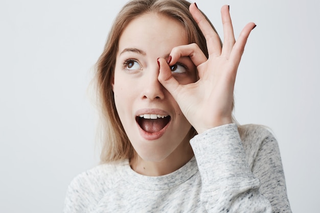 Spaß, Freude und Glück. Bild einer fröhlichen glücklichen jungen Frau mit langen blonden Haaren mit geöffnetem Mund, die mit der Hand eine gute Geste macht, sich über einen guten Tag freut und zeigt, wie großartig sie ist