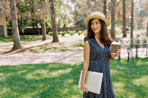 Spektakuläre, charmante Dame mit dunklem Haar, Kleid und Hut hält Laptop und Kaffee im Park