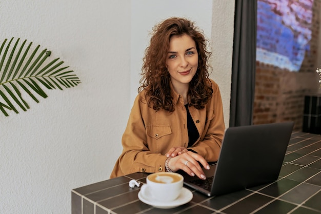 Spektakuläre charmante Dame mit Locken in braunem Hemd, die im Café mit Kaffee sitzt und an sonnigen, warmen Tagen an der Laptop-Fernbedienung arbeitet