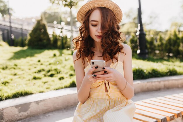 Spektakuläre junge Dame in der trendigen Hut-SMS-Nachricht beim Sitzen im schönen Park. Foto im Freien von modischem Mädchen mit Ingwerhaar, das jemanden auf Bank wartet.