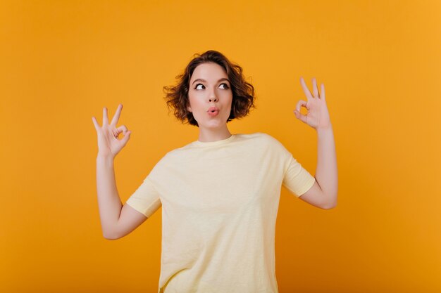 Spektakuläre junge Frau mit Tätowierung lustiges Tanzen auf heller Wand. kurzhaariges sorgloses Mädchen trägt gelbes T-Shirt.