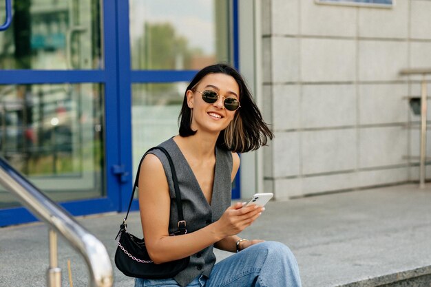 Spektakuläre schöne Frau mit Kurzhaarfrisur in Sonnenbrille mit Tasche hält Smartphone und blickt mit schönem Lächeln in die Kamera