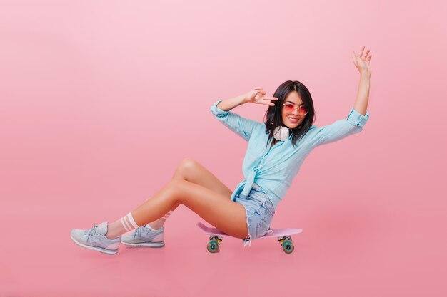 Spektakuläres lateinamerikanisches Mädchen im niedlichen Outfit, das auf Skateboard sitzt und Hände mit glücklichem Gesichtsausdruck winkt. lachende hispanische junge Frau in Sonnenbrille und trendigen Turnschuhen.