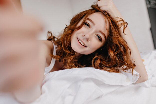 Spektakuläres weißes Mädchen mit Ingwerhaar, das sich am Morgen entspannt. Innenfoto der reizenden kaukasischen jungen Dame, die selfie im Bett macht.