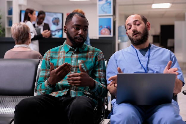 Spezialist, der einen afroamerikanischen Mann im Wartebereich berät und mit einem Laptop die Krankheitsdiagnose und Genesungsbehandlung in der Empfangslobby erklärt. Leute, die in der Einrichtung über Gesundheitsversorgung sprechen.