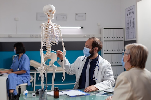 Spezialist erklärt einer pensionierten Frau bei einem Kontrollbesuch das menschliche Skelett und spricht über Knochenverletzungen bei einem Osteopathie-Termin während einer Covid-19-Pandemie. Arzt zeigt Patienten Gelenkmodell.