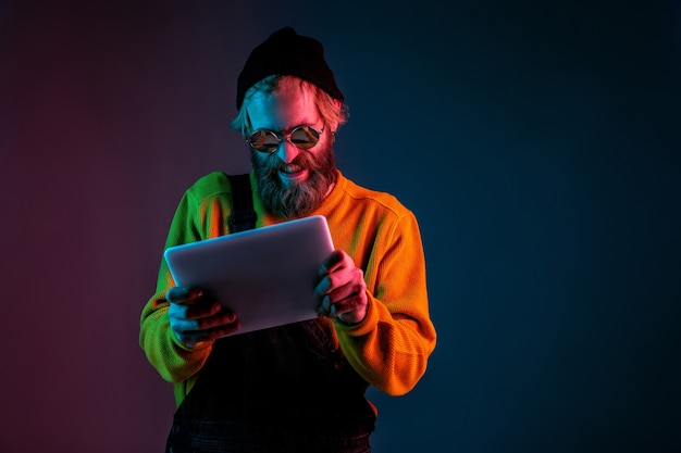 Spiel mit dem Tablet spielen. Porträt des kaukasischen Mannes auf Gradientenstudiohintergrund im Neonlicht. Schönes männliches Modell mit Hipster-Stil. Konzept der menschlichen Emotionen, Gesichtsausdruck, Verkauf, Anzeige.