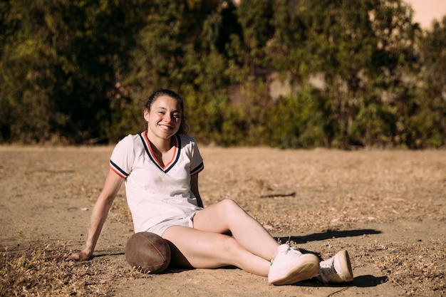 Spielerische junge Frau, die mit Rugbyball sitzt