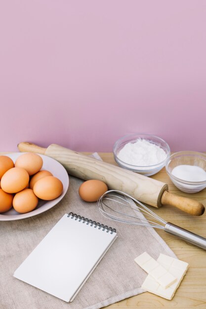 Spiralblock Eier; Schneebesen; Nudelholz; Zucker; weiße Schokolade und Mehl auf dem Schreibtisch