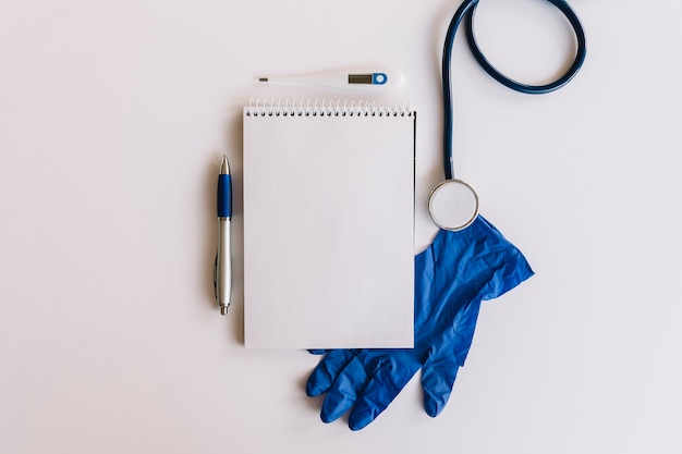 Spiralblock Stift; Stethoskop; Thermometer und Handschuhe auf weißer Oberfläche