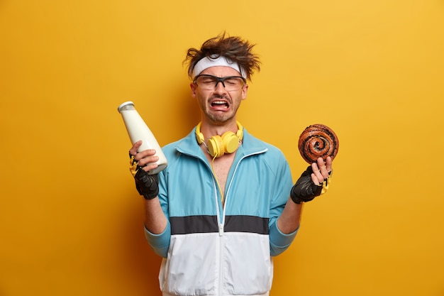 Kostenloses Foto sport-, gewichtsverlust- und versuchungskonzept. emotional missfallener mann in sportbekleidung, hält eine flasche milch und ein köstliches süßes brötchen, ist zuckersüchtig, macht sport, um fit und gesund zu bleiben