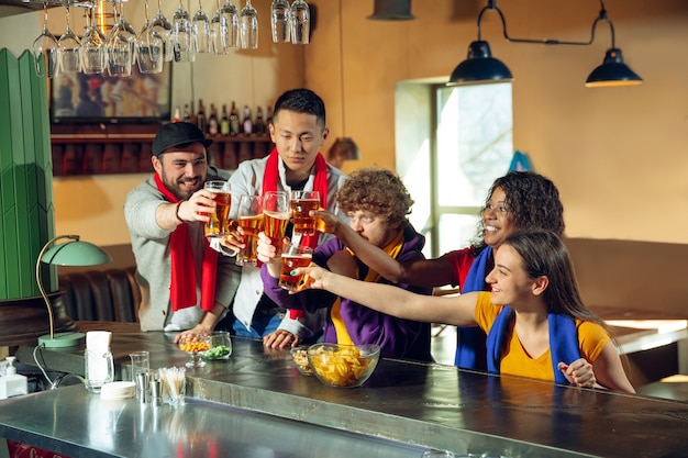 Sportfans jubeln an der Bar, im Pub und trinken Bier während der Meisterschaft, der Wettbewerb geht. Multiethnische Gruppe von Freunden aufgeregt, die Übersetzung zu sehen. Menschliche Emotionen, Ausdruck, unterstützendes Konzept.